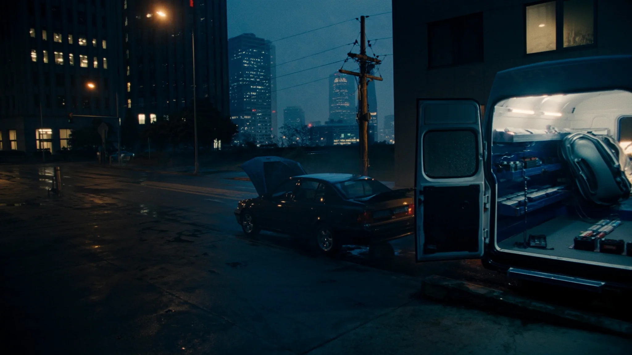 A mobile battery service van with its rear doors open and its racks lit, parked beside a car with a raised hood on a wet city street at night